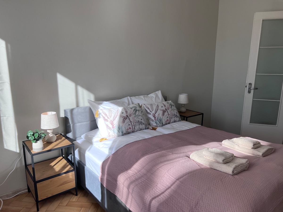 A bright bedroom features a cozy bed dressed with soft linens and decorative pillows. Nightstands with lamps are positioned on either side, complementing the light-colored walls. Natural light floods the space through a nearby window, enhancing the warm, inviting feel of the room.