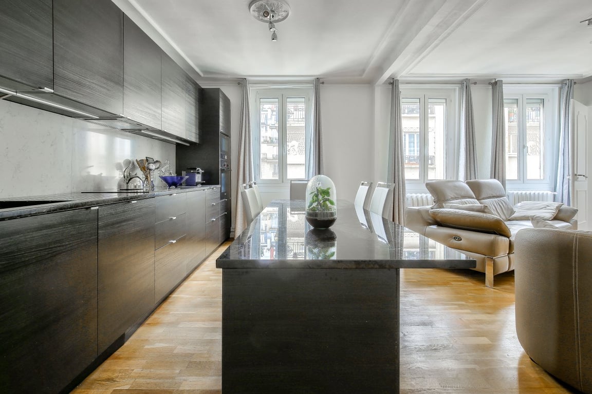 A modern kitchen space is complemented by sleek dark cabinetry and a large island with a polished countertop. The dining area features a round table with comfortable seating. Natural light fills the room through multiple windows, enhancing the airy feel.