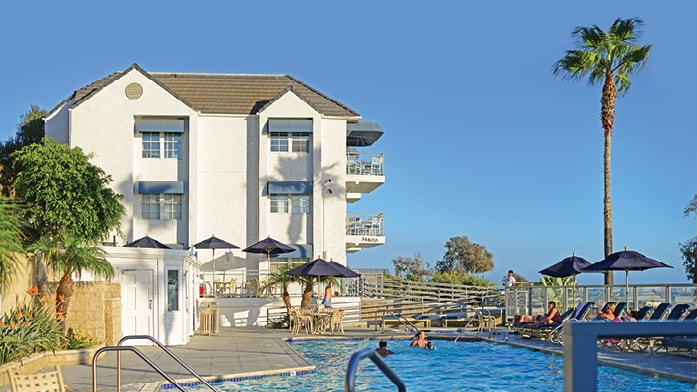 A three-story building with white walls is visible beside a clear outdoor pool. Sun loungers are arranged near the pool area, while umbrellas provide shaded seating. Lush greenery surrounds the space, enhancing a relaxing atmosphere with a view of palm trees and the sky.