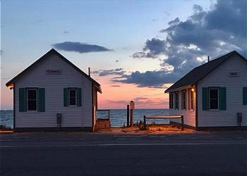 Two classic cottages are positioned along the coast, facing Cape Cod Bay. A soft sunset casts gentle hues over the water, while the cottages' shutters are painted in a calming green. A wooden fence borders the path to the beach, enhancing the seaside ambiance.