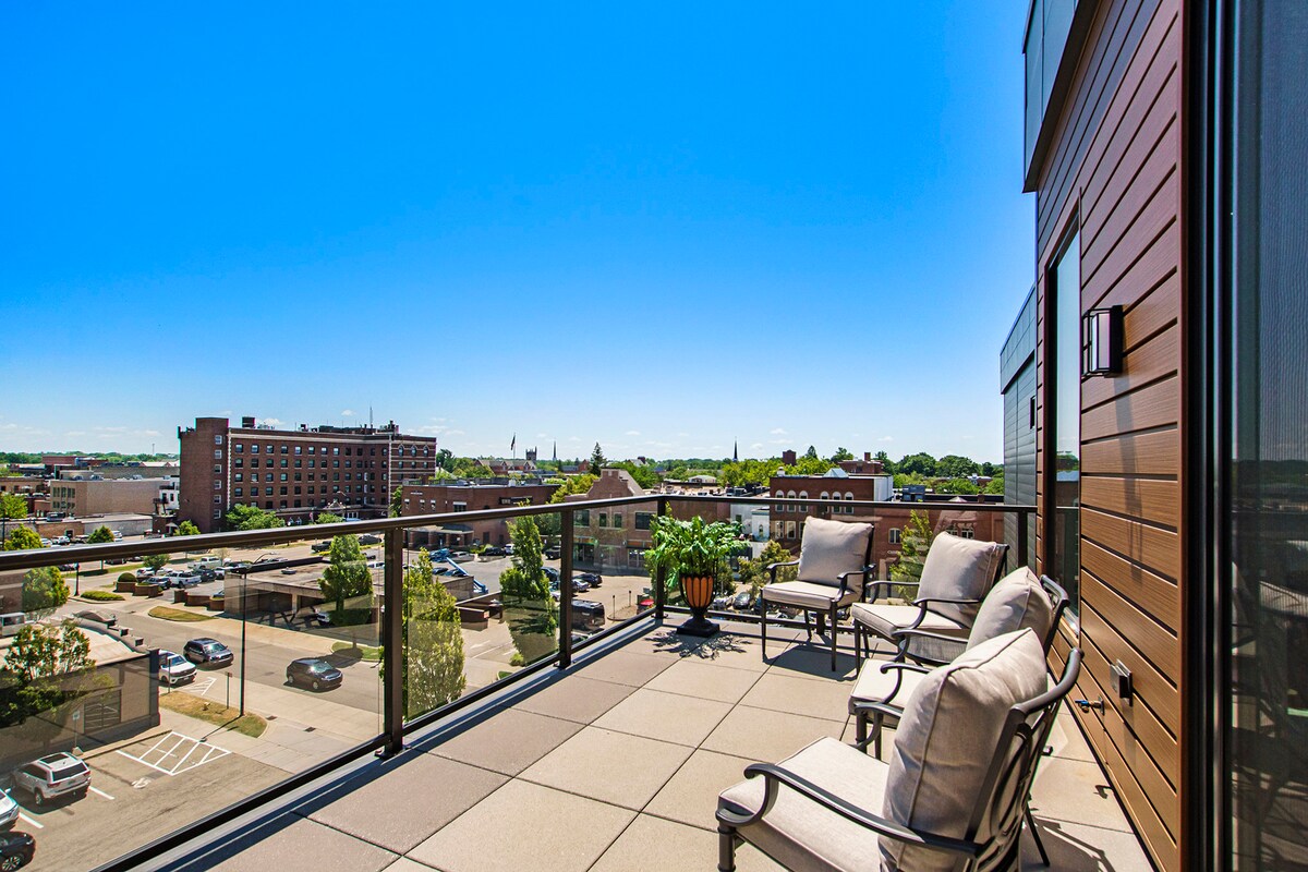 A private balcony is displayed, furnished with comfortable seating arrangements, offering a view of the cityscape below. The surrounding area features a mixture of buildings and green spaces, with clear blue skies above, creating a serene outdoor setting.