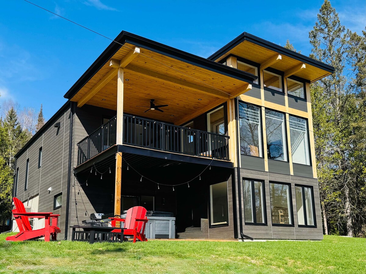 An architectural chalet is showcased with a large balcony supported by wooden beams. Floor-to-ceiling windows reflect the natural surroundings. Red outdoor chairs are arranged around a coffee table on the patio, with a hot tub visible on one side, creating an inviting outdoor space.