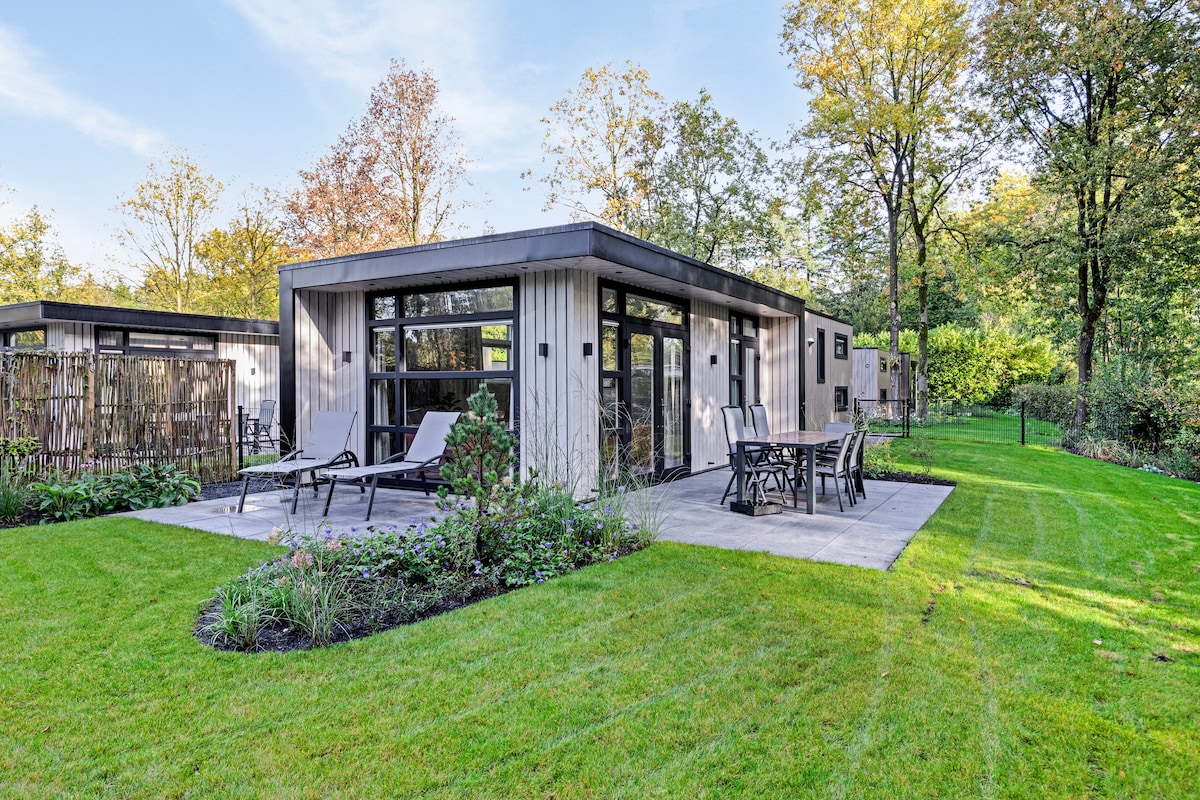 A modern holiday cabin is set amidst lush greenery, featuring large windows that allow natural light. A spacious patio area is furnished with a dining table and lounge chairs, surrounded by manicured lawns and landscaped flower beds.