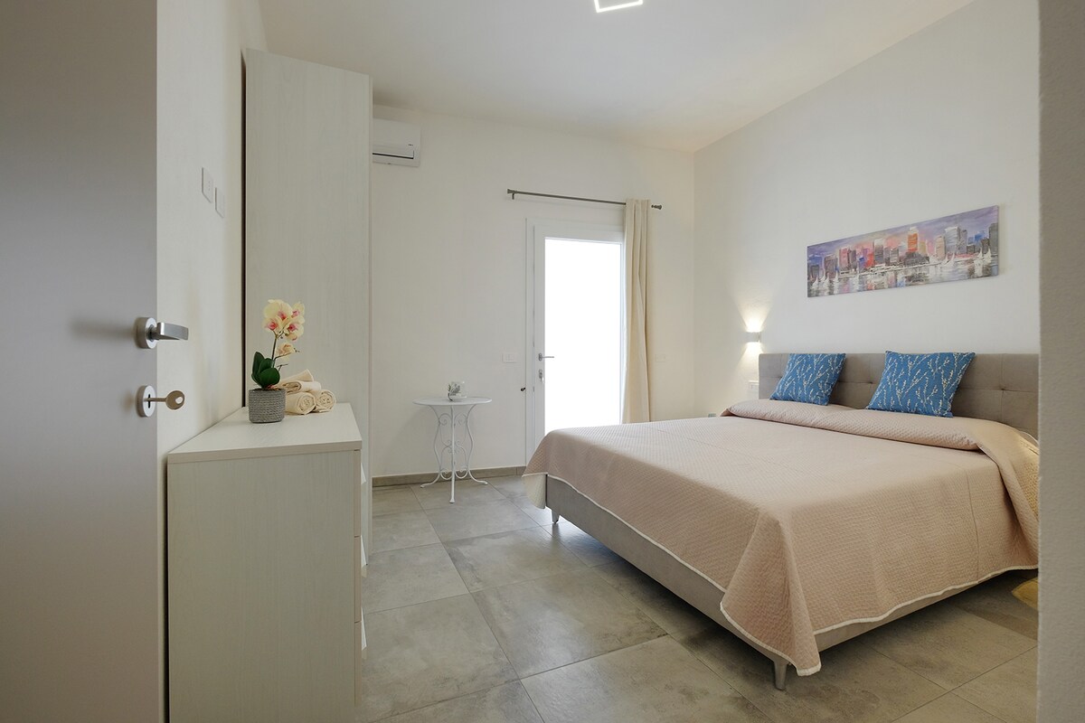 A modern guest room features a spacious bed with soft linens and decorative pillows. Natural light enters through a glass door that leads outside. A small table and chair are positioned near the door, while a dresser adds functionality to the serene space.