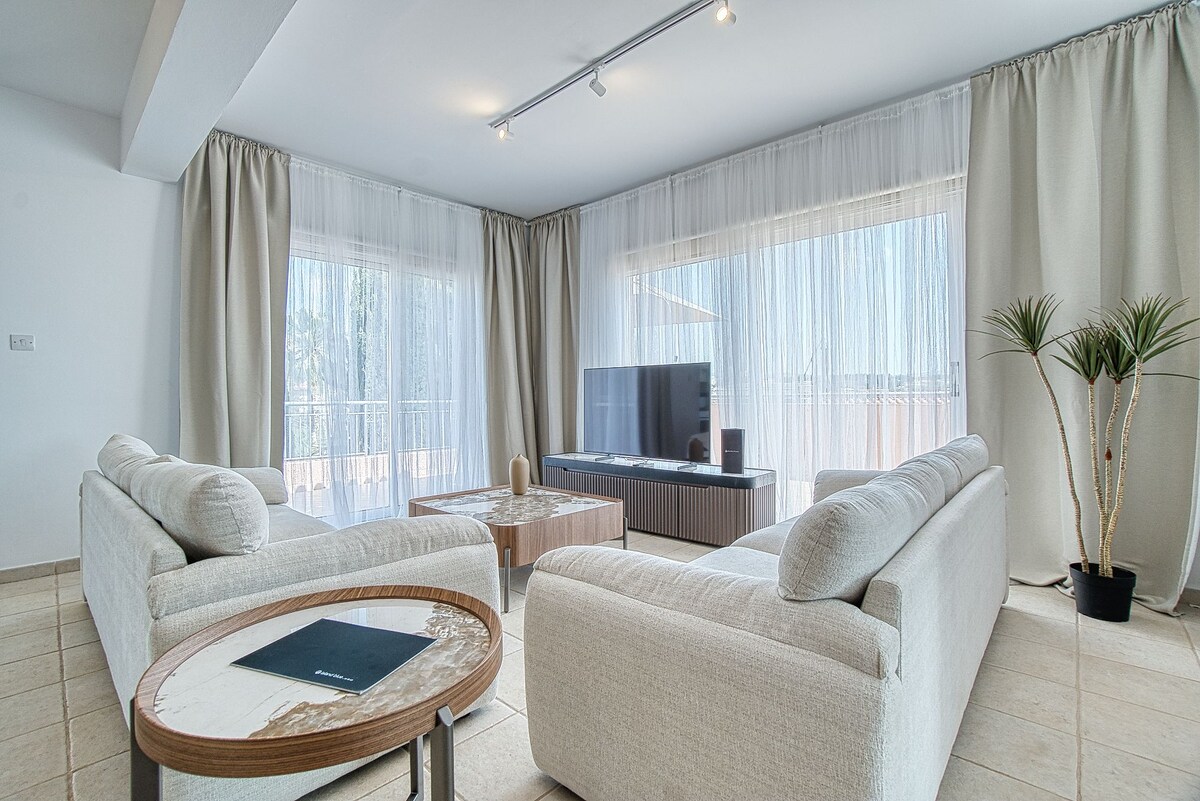 A bright living area features two light-colored sofas positioned around a coffee table. Sheer curtains frame large windows, allowing natural light to fill the space. A modern television is mounted on the wall, and a potted plant adds a touch of greenery to the room.