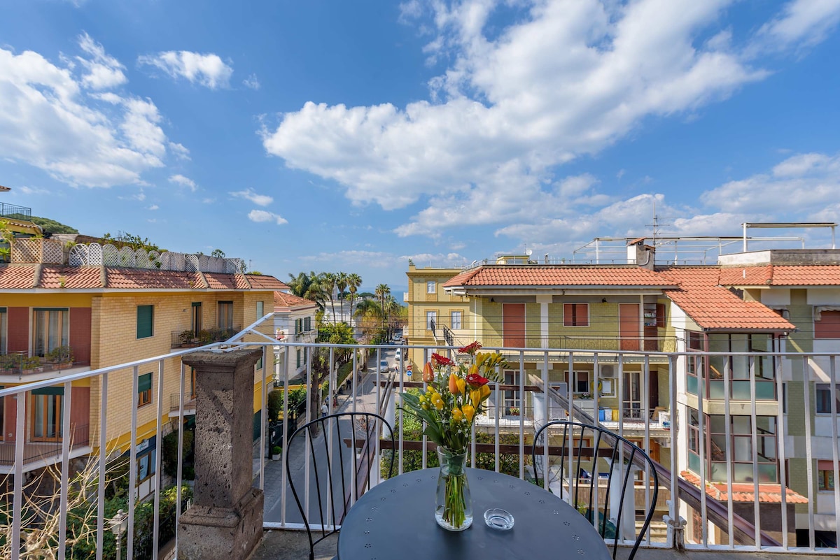 A charming terrace is highlighted by a black table set for two, adorned with a vase of colorful flowers. Views of the neighboring buildings and palm trees under a blue sky with scattered clouds create a pleasant outdoor setting.