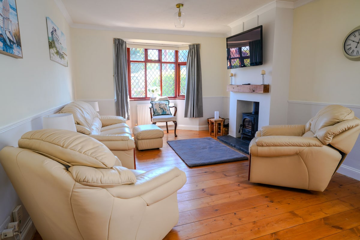 The living area features light-colored leather sofas arranged around a small table, with a cozy rug on the wooden floor. Large windows allow natural light to fill the room, complemented by neutral walls and curtains. A wall-mounted flat-screen TV is visible, alongside a decorative clock.