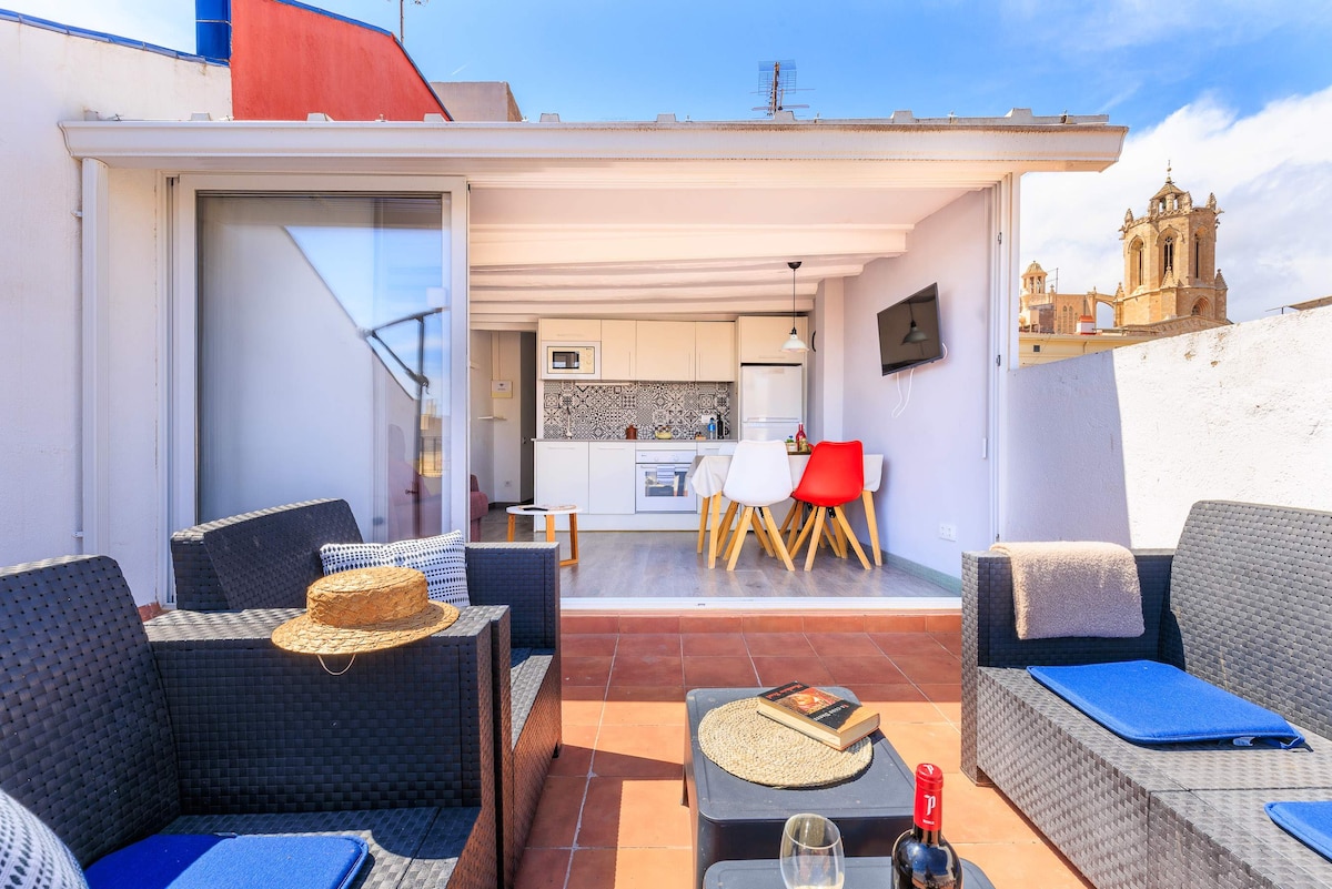 A spacious terrace is presented with comfortable seating arrangements featuring wicker chairs and blue cushions. A modern kitchen area is visible through large glass doors, while a glass of wine and a book are set on the table. Historic buildings are seen in the background.