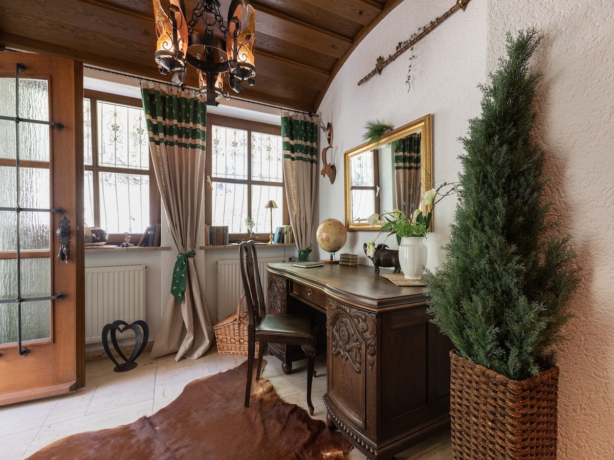 A home office corner displays a wooden writing desk with a globe and decorative plants. Natural light filters through two windows adorned with green-trimmed curtains. A decorative mirror and a potted plant are positioned on the desk, alongside a textured cowhide rug on the floor.
