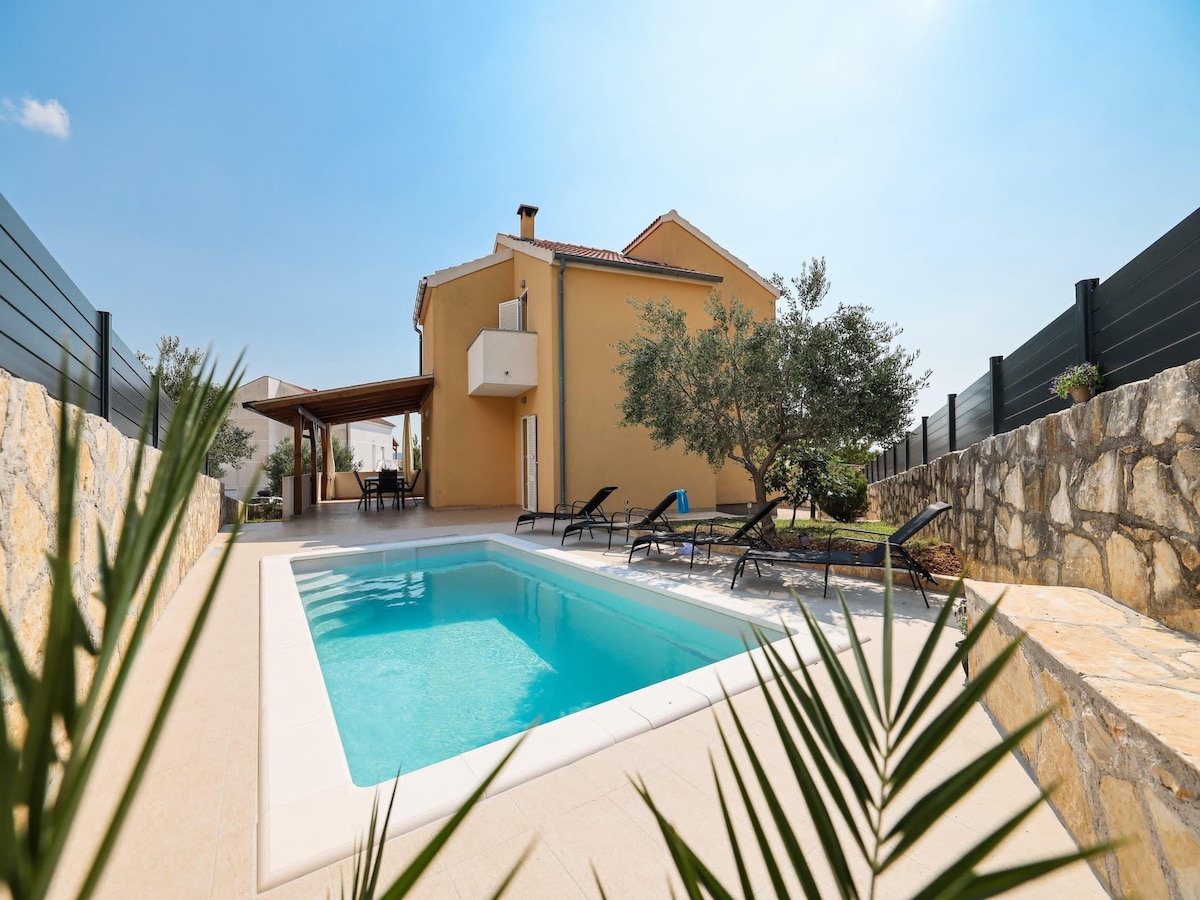 A private swimming pool is framed by stone walls and garden details, creating a serene outdoor space. Sun loungers are arranged nearby for relaxation. The house stands prominently in the background, complemented by greenery and an outdoor terrace area, inviting pleasant gatherings.