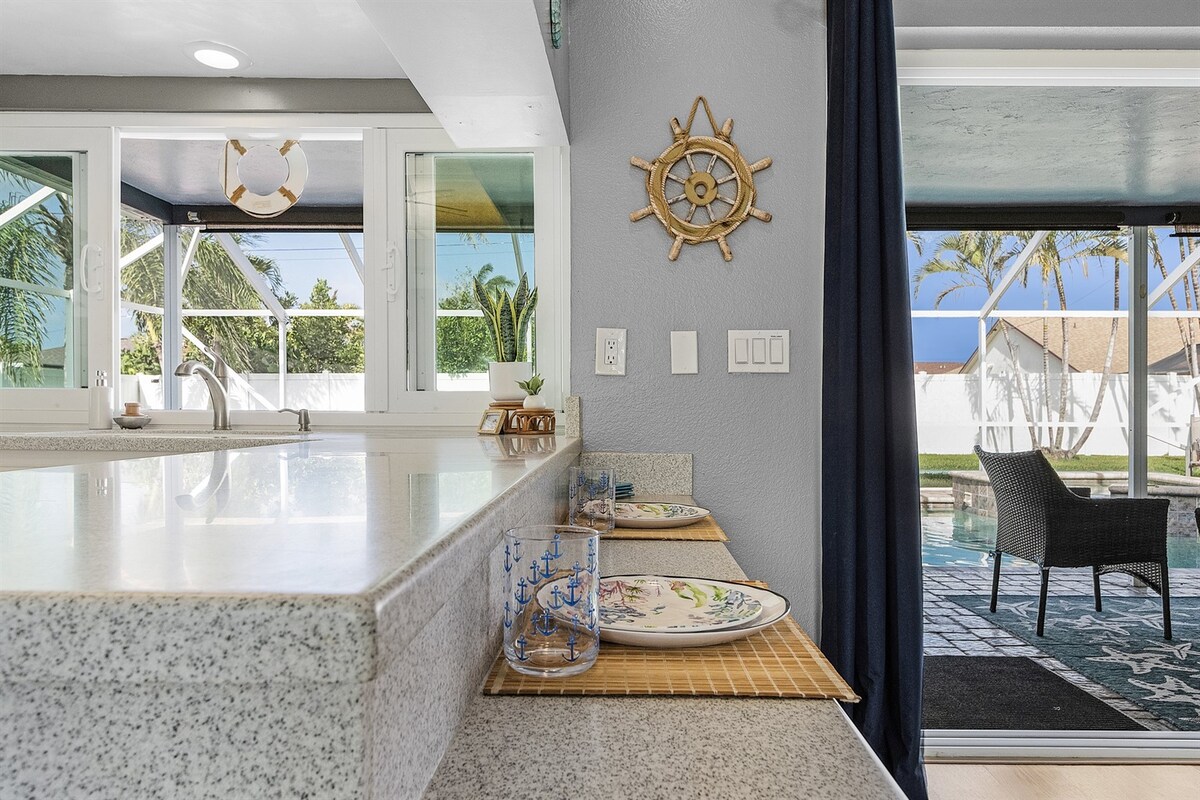 A well-appointed kitchen countertop is visible, featuring two place settings with dishes and glasses arranged neatly. The background reveals large windows that provide a view of the outdoor pool area, where palm trees can be seen swaying gently in the breeze.