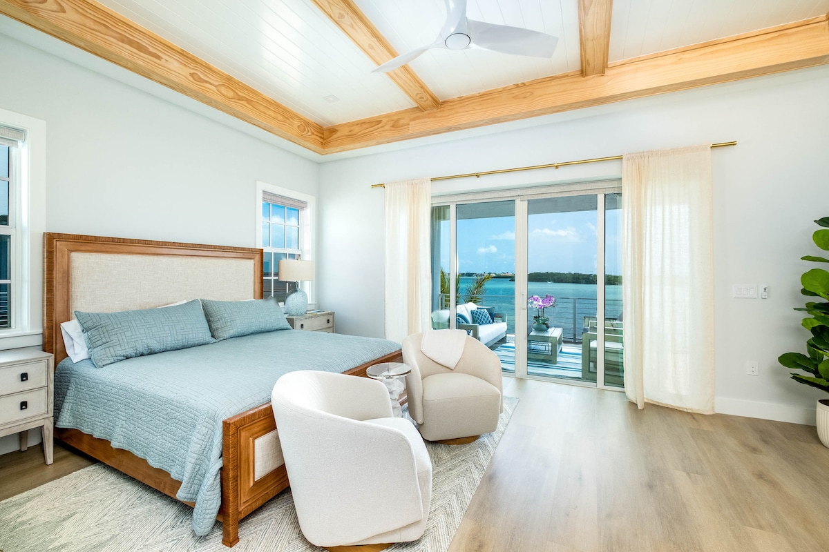 Waterfront king suite in a luxury Marathon Middle Keys villa with private balcony views over the Atlantic, soft coastal design, wood beam ceiling, and lounge seating. Premium Florida Keys lodging in SeaGlass Cove near marinas and beaches.