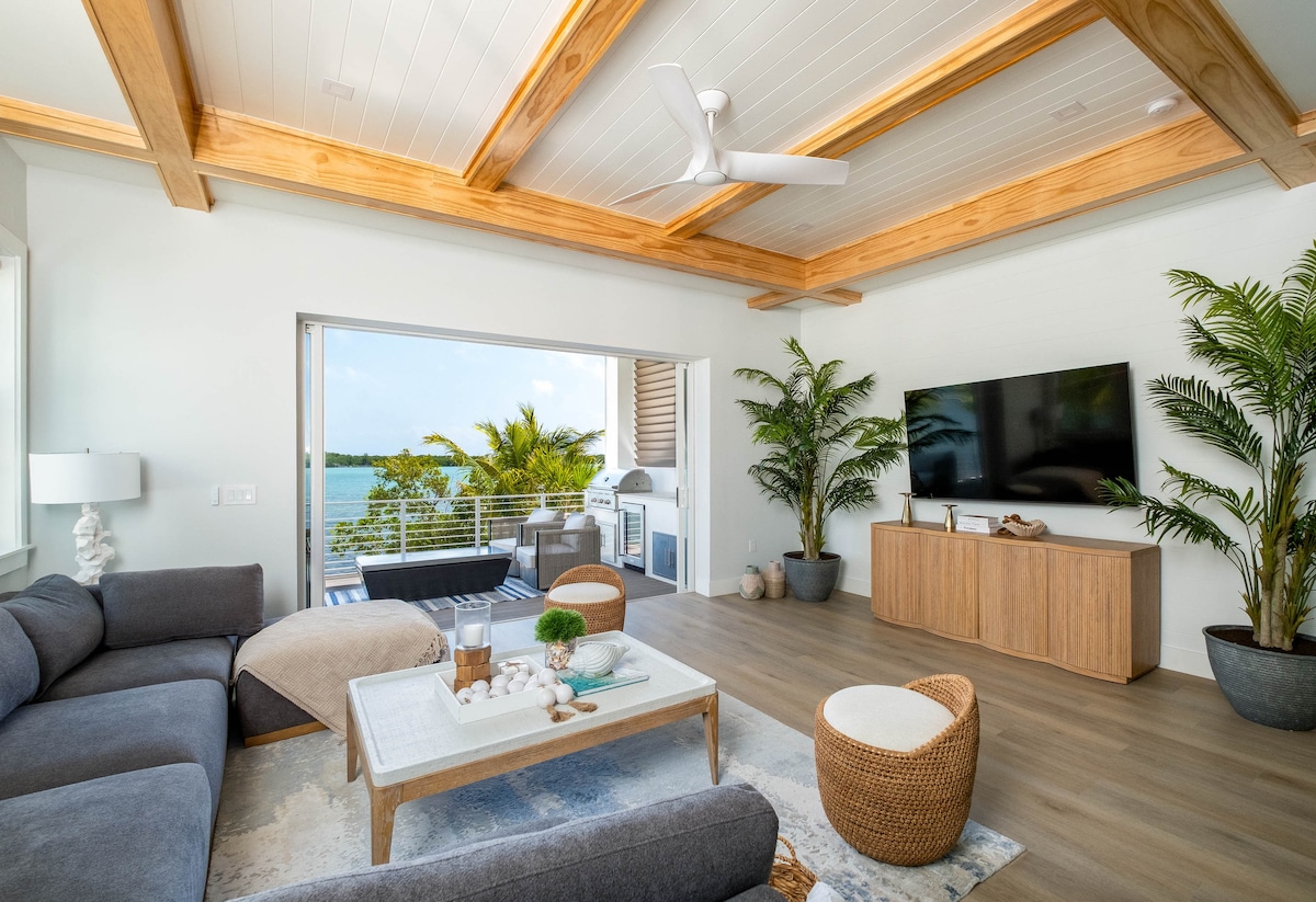 Coastal living room in a luxury Marathon Middle Keys oceanfront home, featuring wood beam ceiling, smart TV, designer furnishings, and sliding doors opening to a waterfront terrace with grill kitchen. 5BR Florida Keys villa in SeaGlass Cove.