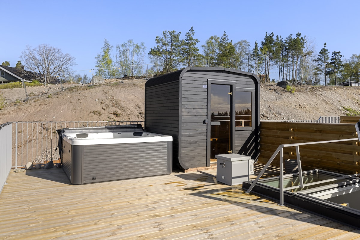 The rooftop terrace features a wooden deck, housing a dark wooden sauna and a round jacuzzi. Surrounded by green trees, this relaxing space provides a tranquil atmosphere. A lounge chair is visible nearby, enhancing the area designed for leisure and enjoyment.
