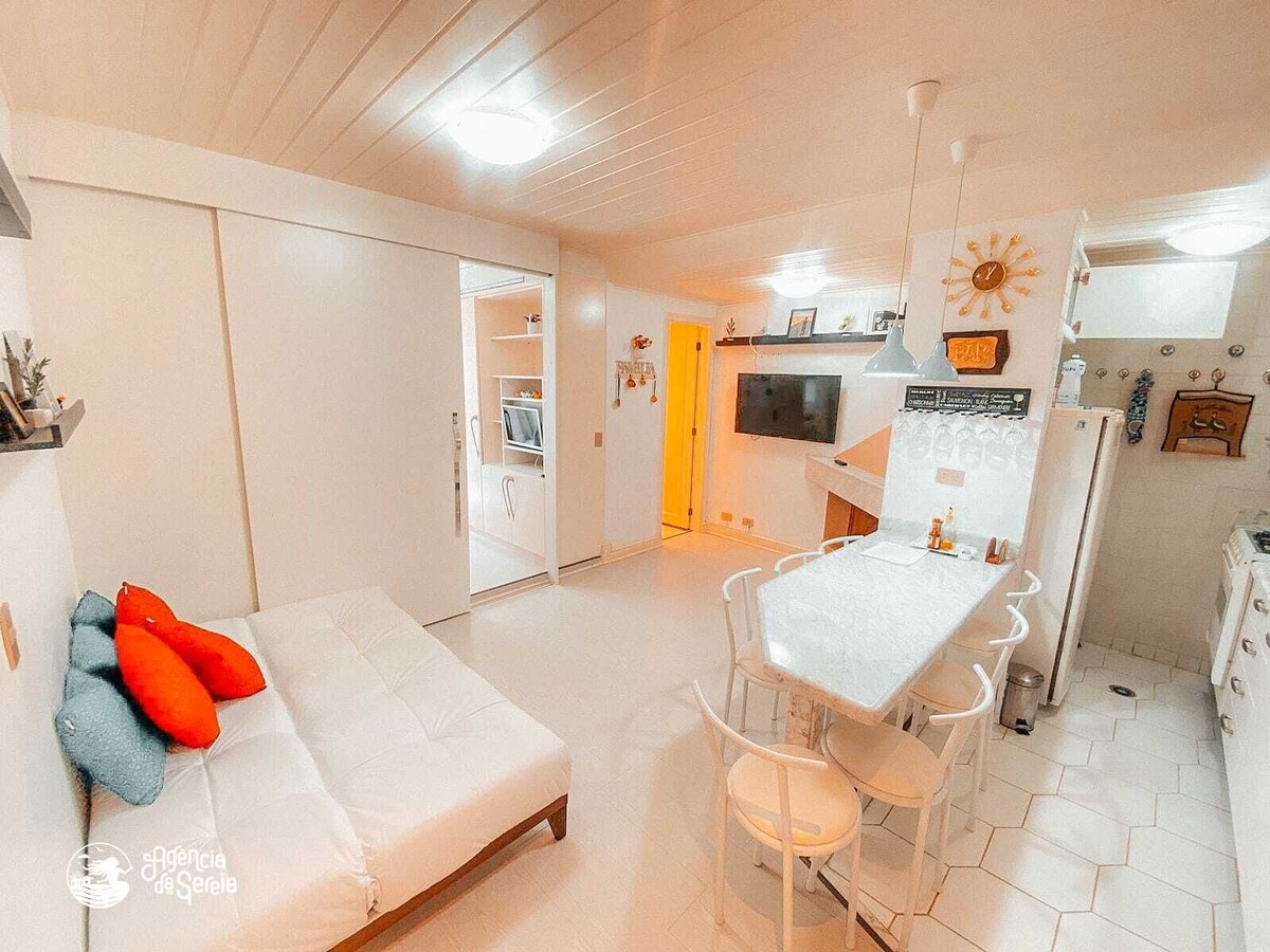 A bright studio space features a white color scheme, with a spacious sofa bed and a dining area equipped with a light-colored table and white chairs. The kitchen is fully equipped, showcasing a refrigerator and stove, while soft lighting contributes to a warm ambiance.