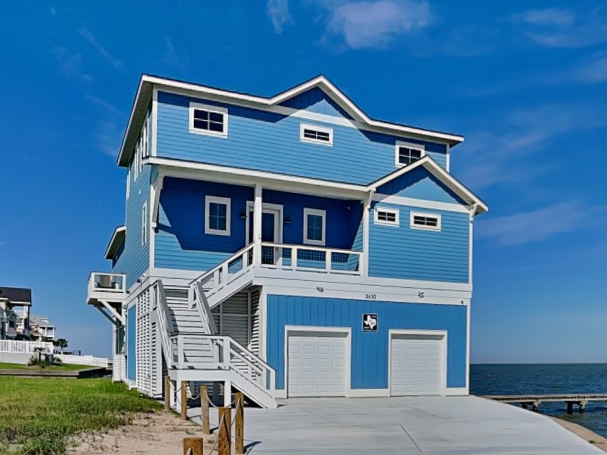 A bright blue three-story house is prominently featured with a spacious driveway and two garage doors. Architectural details include multiple balconies and expansive windows, providing views of the waterfront. The structure is set against a clear blue sky, creating a welcoming coastal vibe.