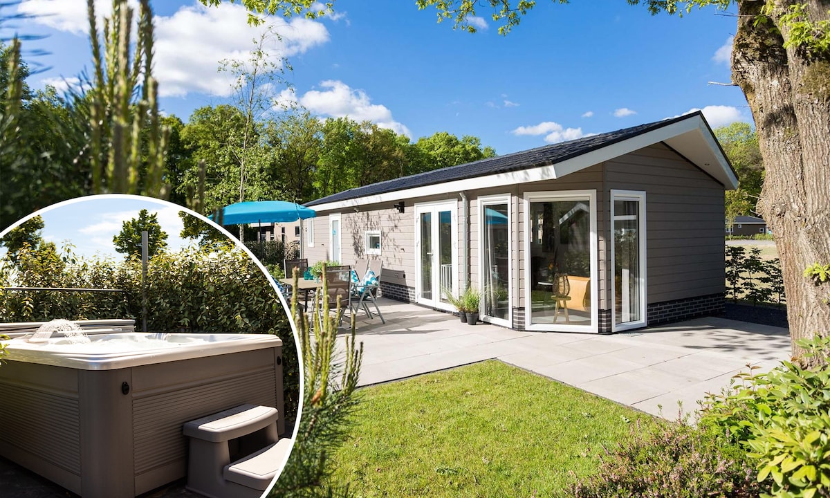 A modern holiday home is set within a green landscape, featuring large glass doors that invite natural light. A private hot tub is positioned on a stone patio, surrounded by manicured grass and shrubbery. Outdoor seating areas are visible under blue umbrellas.