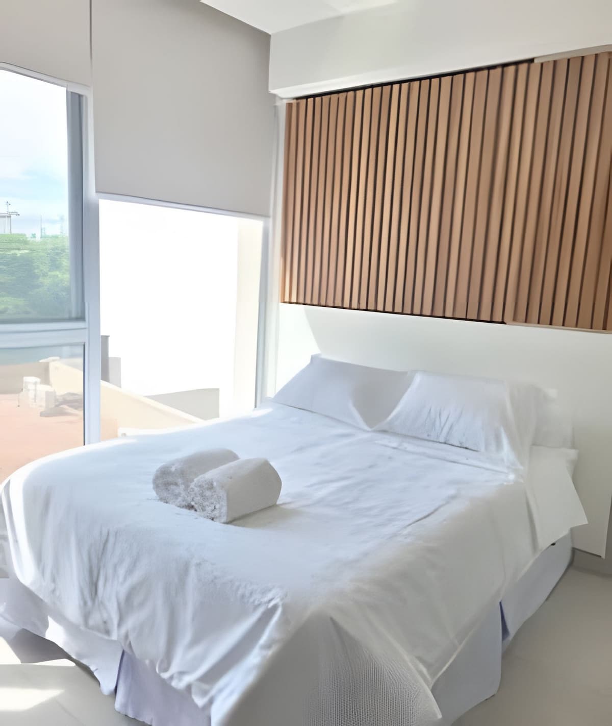 A bright bedroom is presented with a spacious bed covered in a clean white bedspread. A soft towel is neatly placed on the bed. Natural light floods the room through a large window, with textured wood paneling adding visual interest to the wall.
