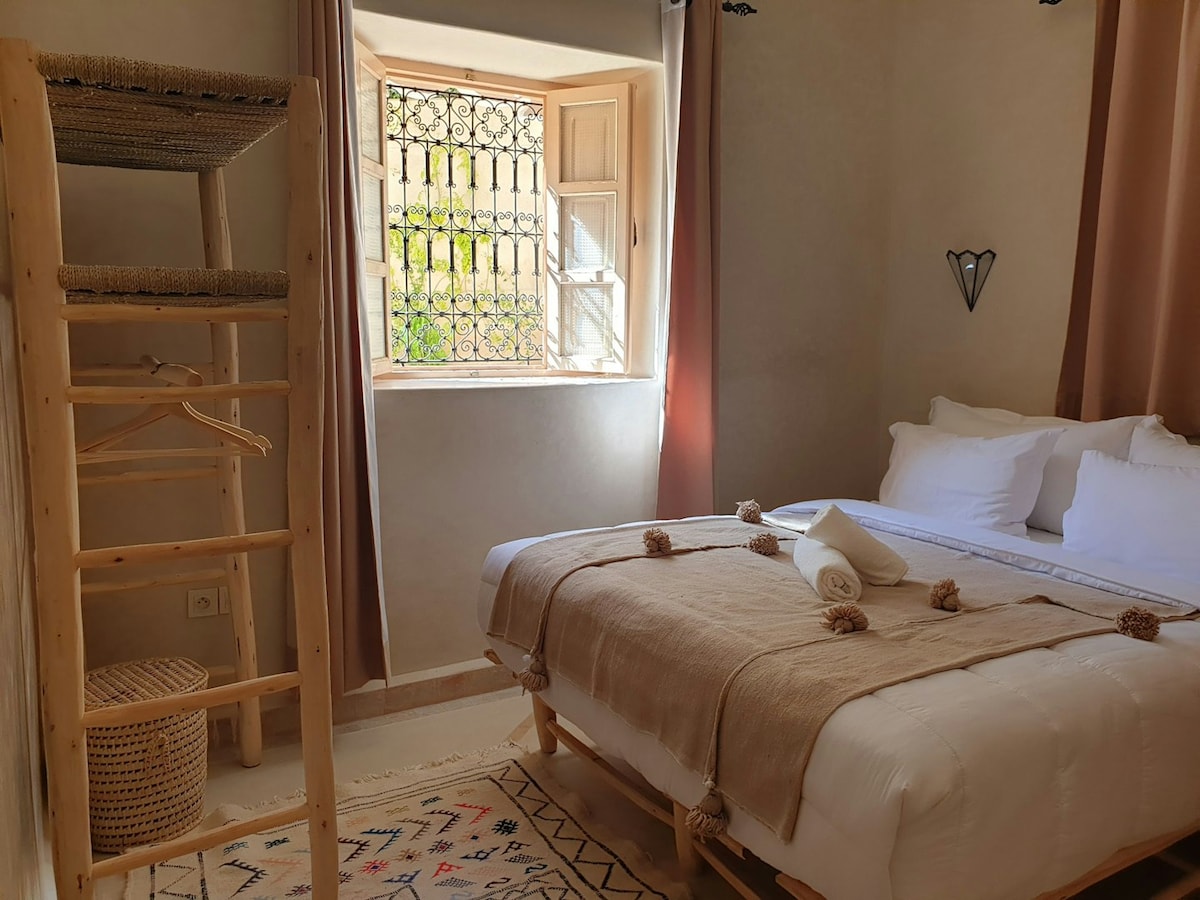 A cozy room features a spacious bed with a neutral-colored blanket, complemented by white bedding. A woven laundry basket is placed nearby, and a wooden ladder shelf stands against the wall. Natural light filters through a decorative window, illuminating the textured decor.