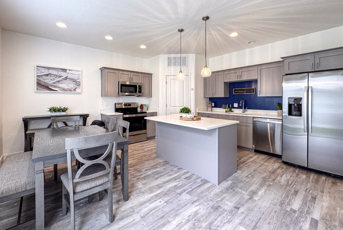 A modern kitchen area is highlighted by stainless steel appliances and quartz countertops. A dining table with seating for six is positioned adjacent to a kitchen island with additional seating. The space is illuminated by pendant lighting, enhancing its contemporary design.