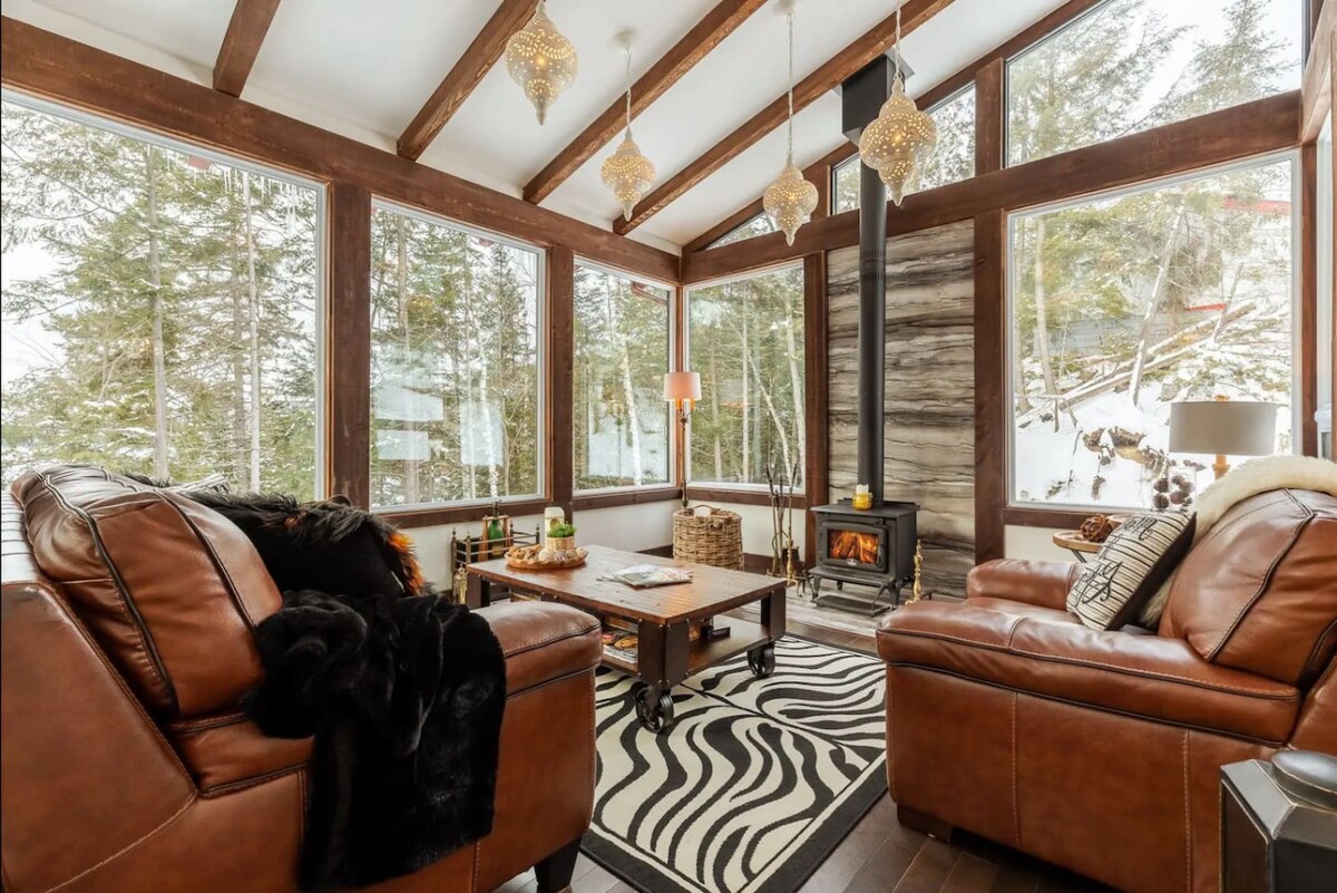 A cozy seating area is presented with rich brown leather sofas, paired with a wooden coffee table. Large windows allow ample natural light, revealing views of the snowy outdoor landscape. Unique light fixtures hang from the ceiling, adding to the inviting ambiance.