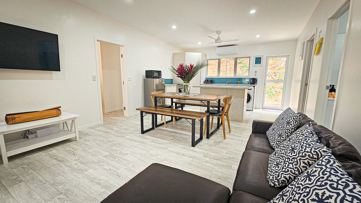 A bright living area features a spacious brown sofa adorned with patterned cushions. A large wooden table with seating occupies the center, while a fully equipped kitchen is visible in the background. Large windows allow natural light to illuminate the space.