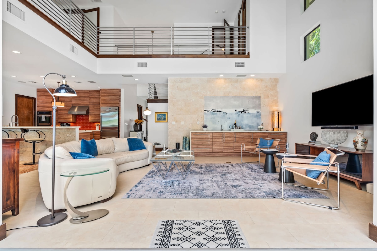 An airy living space features vaulted ceilings and a blend of modern decor. A plush sofa is arranged alongside stylish armchairs and a glass coffee table, set atop a patterned area rug. A large television is mounted on the wall, and a wall of windows allows natural light to fill the room.