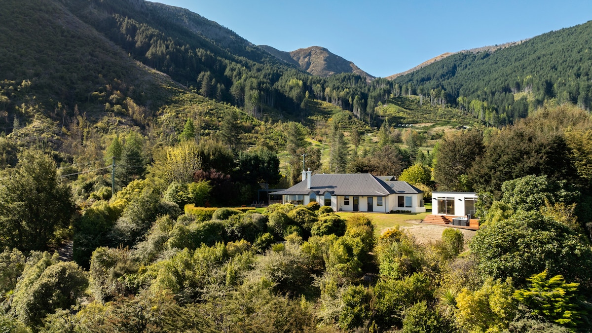 A spacious retreat is set against a backdrop of rolling green hills and forested mountains. The single-story house features a mix of greenery and landscaped gardens, creating a peaceful and inviting environment. The sunlit exterior highlights the tranquil surroundings.