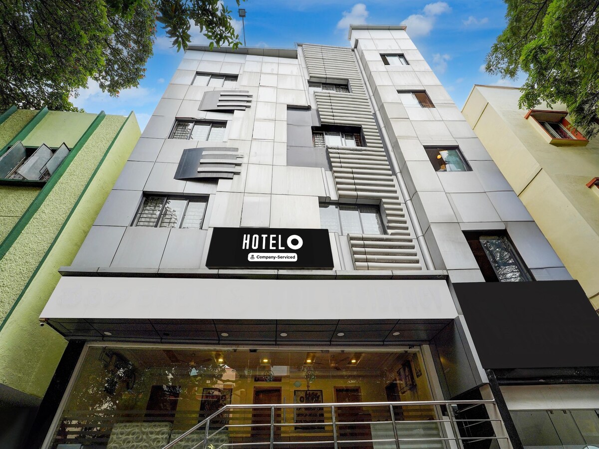 The exterior of Super Hotel O Thimmaiah Rd Rajajinagar is presented, showcasing a modern building design with a sleek façade. Large windows and a prominent hotel sign are visible. A welcoming entrance with steps leads to the main entrance, surrounded by greenery.