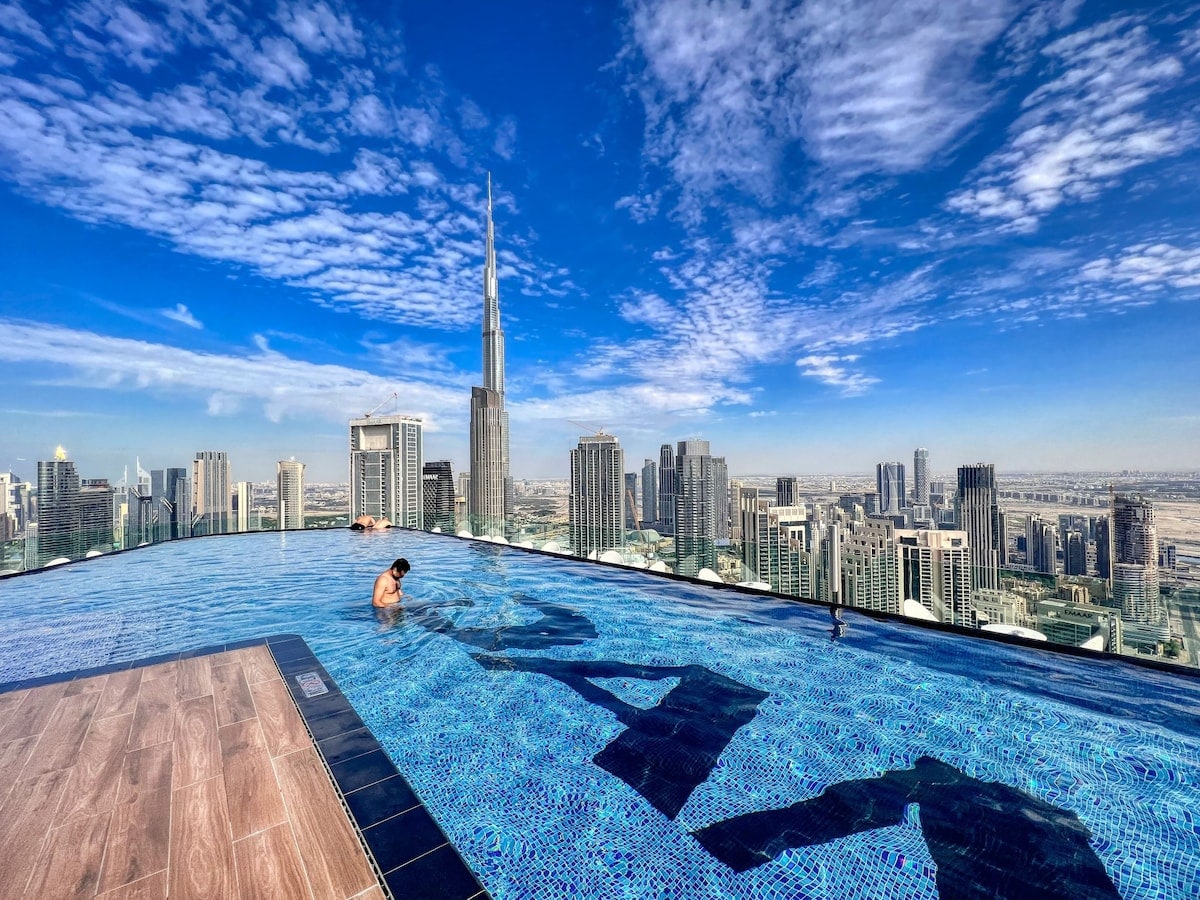 An infinity pool at the 51st floor offers panoramic views of the Dubai skyline, including the Burj Khalifa. The pool features sparkling blue tiles, with a person enjoying the water, and expansive cityscapes stretching towards the horizon under a clear blue sky.
