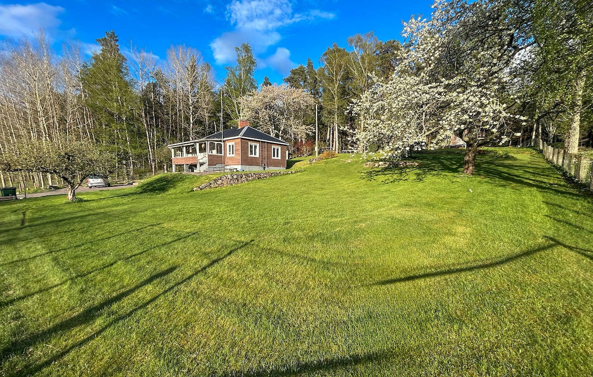 A charming holiday home is set in a spacious garden, surrounded by trees and greenery. The house features large windows, and a pathway leads from the house to a serene lawn area. Lush grass lends a fresh appearance, inviting outdoor activities.
