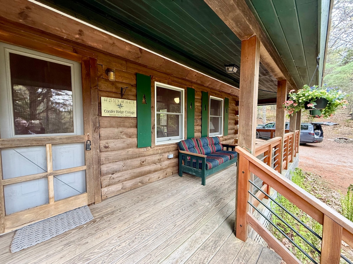 A welcoming entrance leads to a wooden porch featuring a cozy seating area with colorful cushions. The cabin's exterior is constructed from logs, complemented by green shutters on the windows. A sign indicates the property's name, and a vehicle can be seen parked nearby.