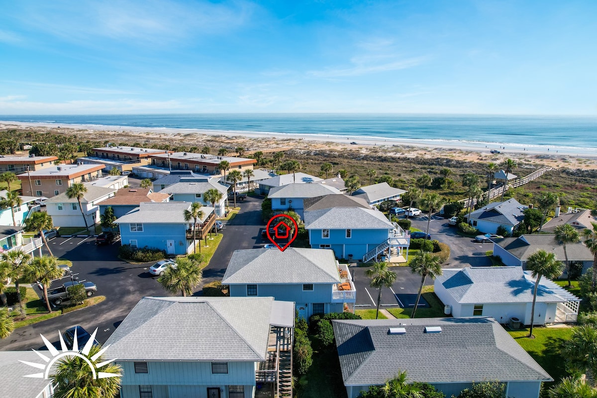 Aerial view of the Surf Crest Village community showcasing a cluster of coastal cottages. The sandy beach stretches along the horizon, with the ocean visible in the background under a clear blue sky. The image captures the inviting layout of the neighborhood.