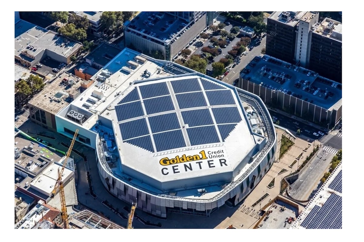 Aerial view of the Golden 1 Center features a uniquely shaped roof equipped with solar panels. Situated in a downtown area, modern buildings are visible in the surrounding landscape. The structure prominently displays the name 'Golden 1 Center' against a bright background.