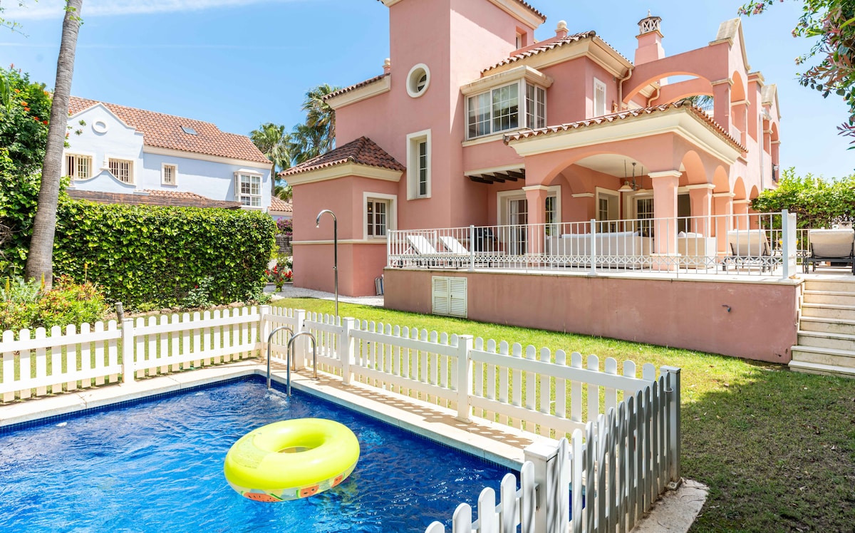 An inviting private pool is surrounded by a white picket fence, featuring a bright yellow pool float. The villa's charming pink exterior and spacious patio areas are visible in the background, framed by lush greenery and a clear blue sky.