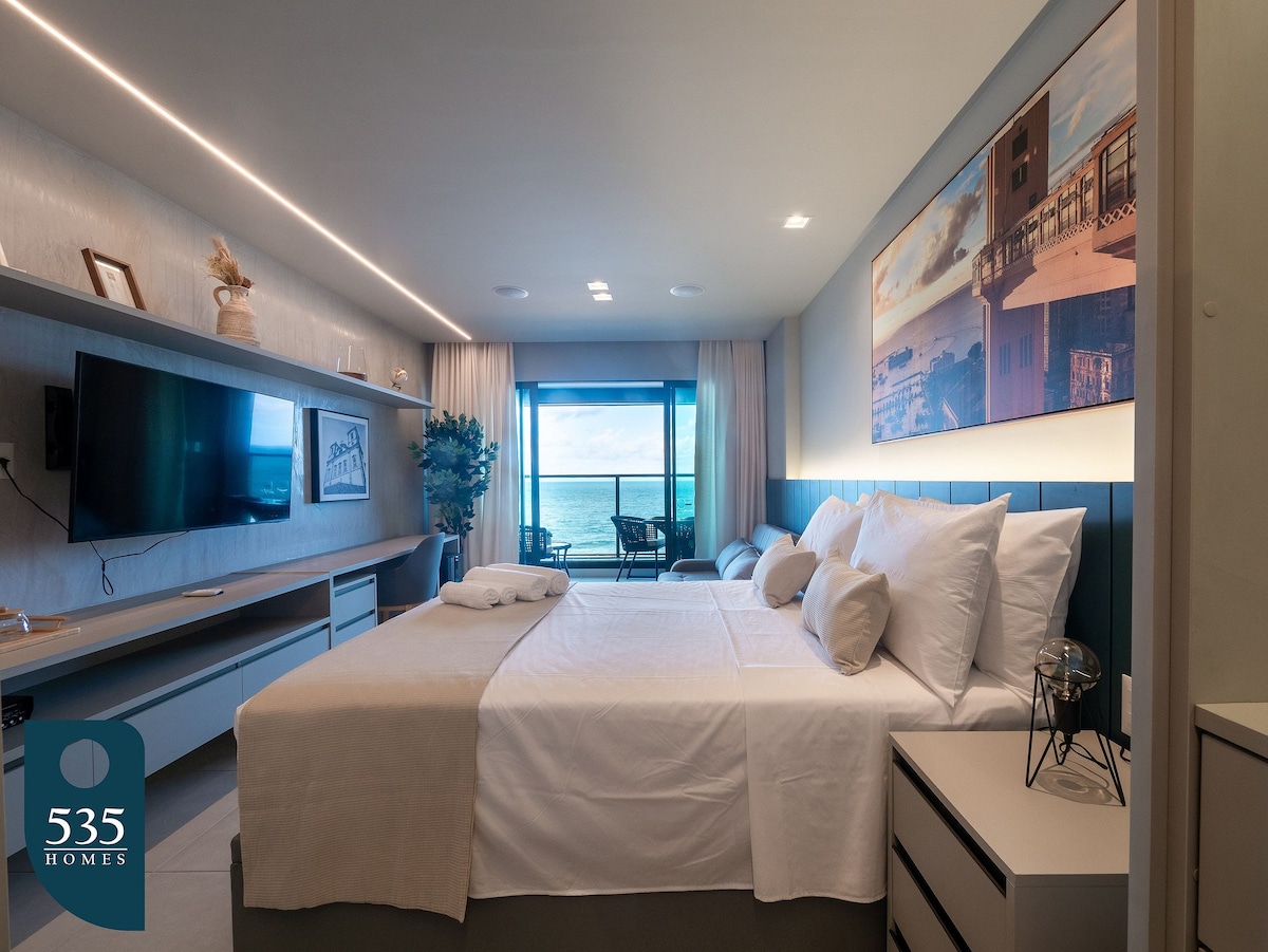A cozy bedroom features a spacious bed with light-colored linens, complemented by soft pillows. A large window showcases an ocean view, while modern shelving and a stylish wall art piece enhance the room’s aesthetic. Natural light illuminates the space, creating a tranquil environment.