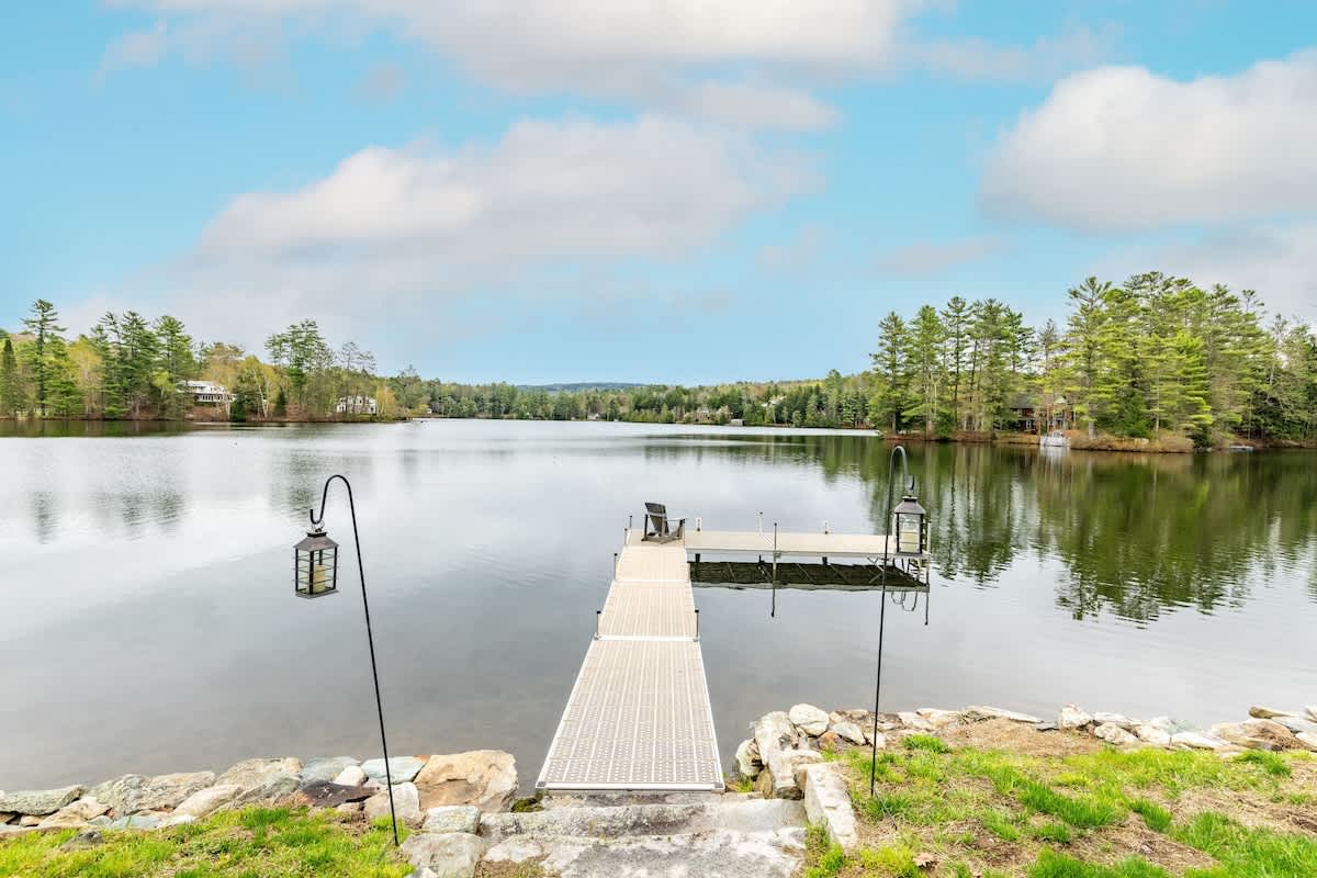 A wooden dock extends over a calm lake, surrounded by trees reflecting on the water's surface. Two lanterns are positioned on either side of the dock. The serene landscape invites relaxation, with gentle skies in the background enhancing the natural setting.