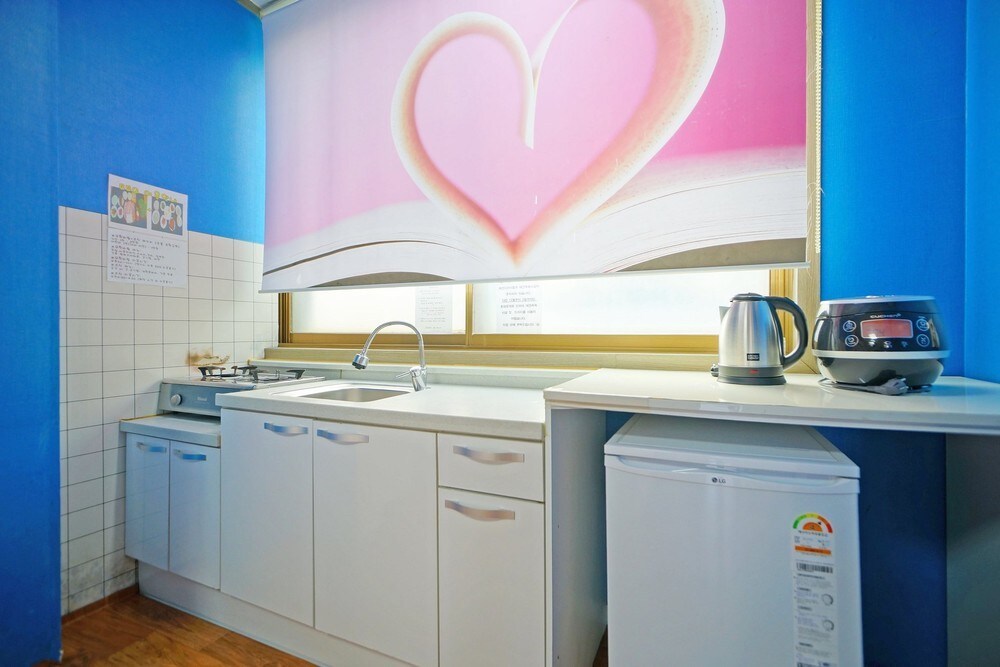 The kitchen area features a blue wall with a heart-themed curtain. A stainless steel sink and countertop are visible, along with a compact refrigerator and an electric kettle. Ample cabinet storage is provided beneath the counter, enhancing functionality.
