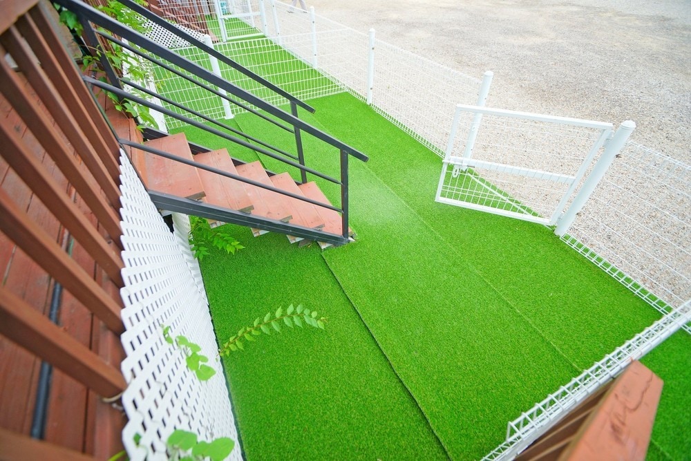 An outdoor space features a green artificial lawn bordered by a white fence. A staircase leads from the upper level to this area, providing easy access. The surrounding environment is designed to be pet-friendly, facilitating outdoor relaxation.