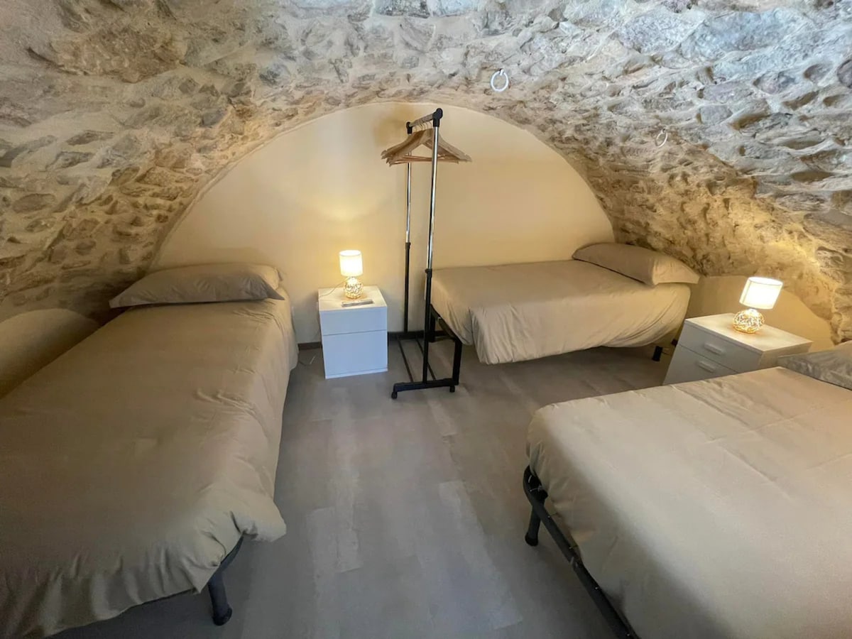 A cozy sleeping area features three spacious beds, positioned beneath a naturally arched stone ceiling. Nightstands with lamps are placed beside the beds, providing soft lighting. The room showcases a warm and neutral color palette, creating a tranquil environment.