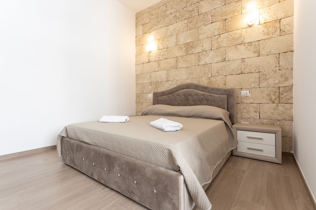 A spacious room features a double bed with a soft, neutral-colored coverlet and two neatly arranged towels. The bed is framed by a textured stone wall, providing a modern ambiance. A bedside table stands next to the bed, with a soft glow emanating from wall-mounted lighting.