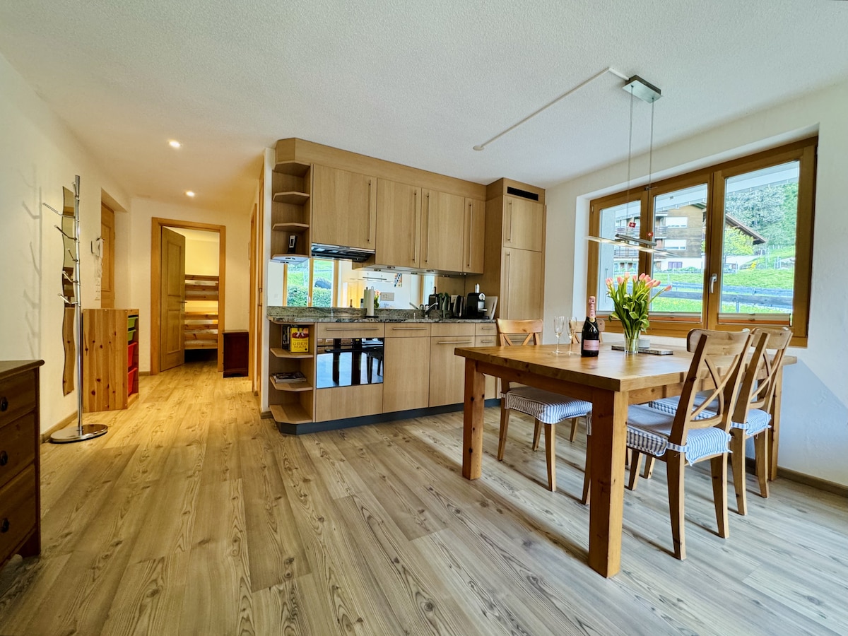A modern kitchen and dining area is presented with light wooden cabinetry. A spacious table is surrounded by four chairs, and natural light flows through the large window, highlighting the warm tones of the wood flooring.