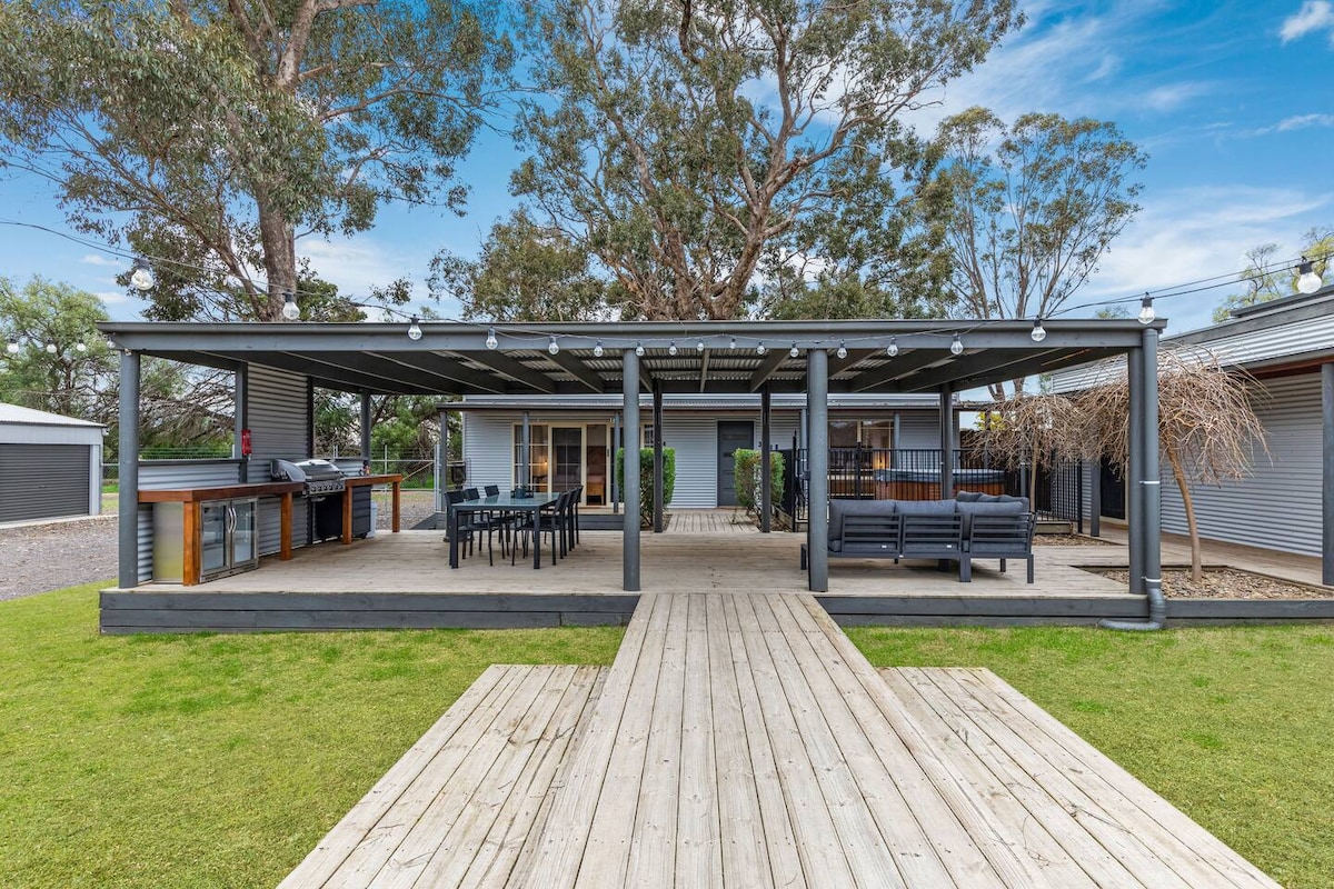 A spacious outdoor area is surrounded by greenery, featuring a large deck with seating and a dining area. A covered patio provides shade, while a barbecue is positioned nearby. The layout is open and welcoming, allowing for relaxation and gatherings.