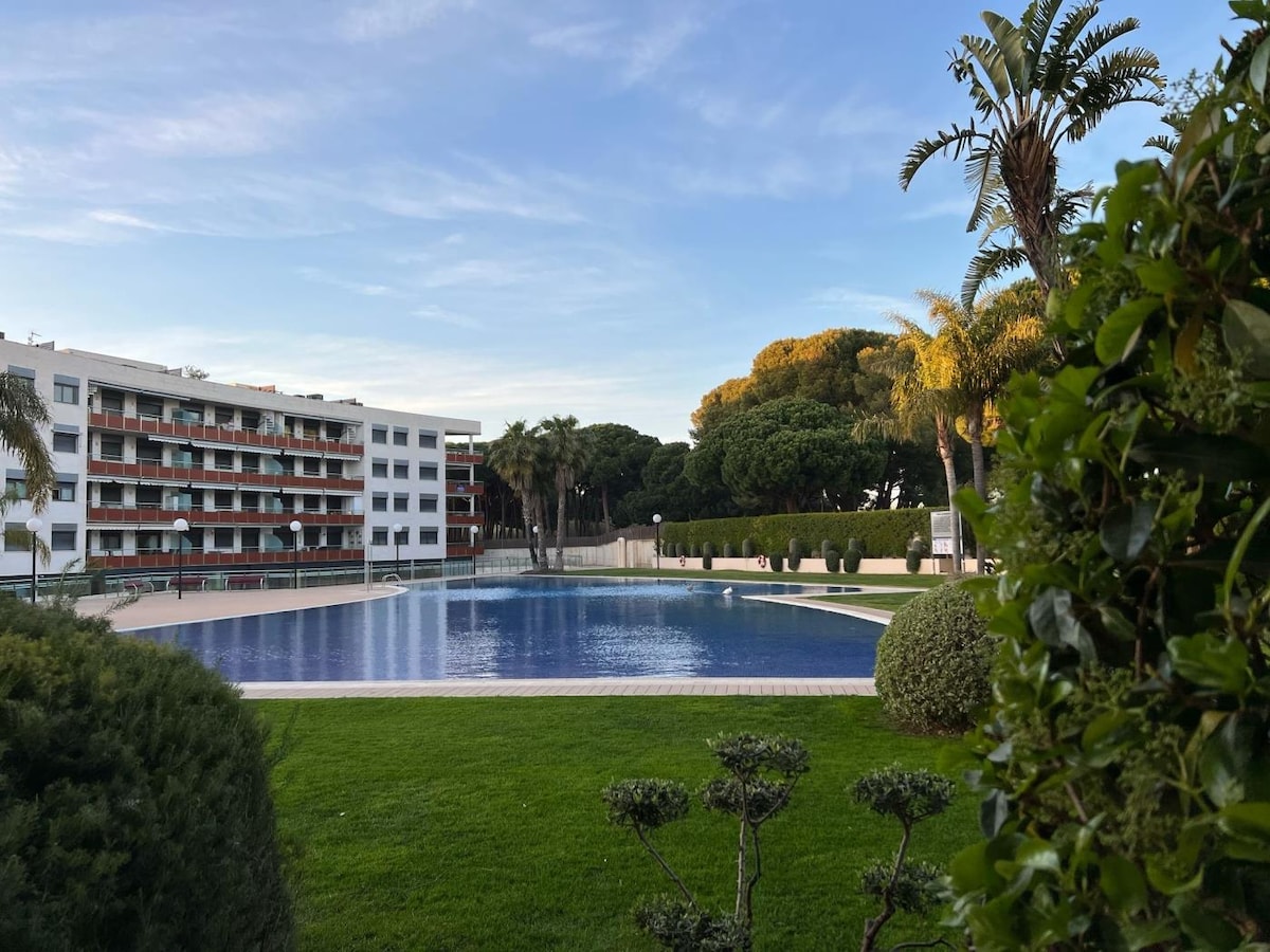 A serene outdoor area is presented with a large, inviting swimming pool surrounded by manicured grass and palm trees. Lush greenery frames the scene, with a modern building visible in the background, highlighting the tranquil atmosphere of the complex.