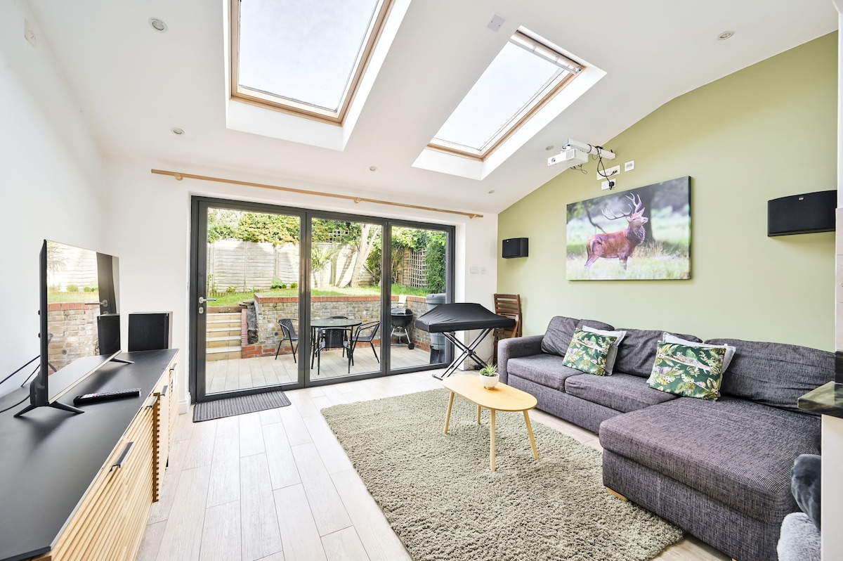 A spacious living area is illuminated by natural light from skylights and large glass doors leading to a garden view. A dark sectional sofa with patterned cushions is complemented by a light wooden coffee table. A wall-mounted TV and art piece enhance the welcoming ambiance.