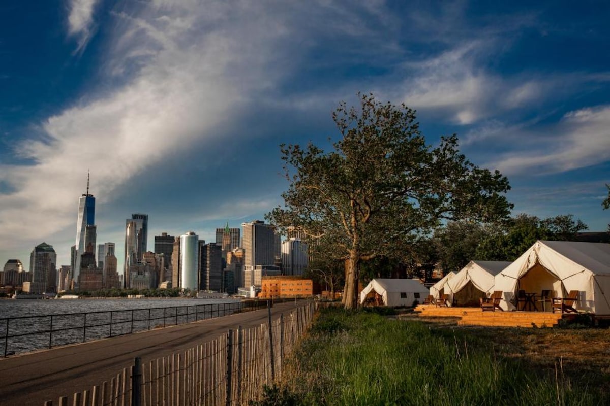 Canvas tents are positioned along a scenic waterfront, with a backdrop of the New York City skyline. Sunlight casts a warm glow, highlighting lush greenery and a large tree near the tents. The calm water reflects the towering buildings under a partially cloudy sky.
