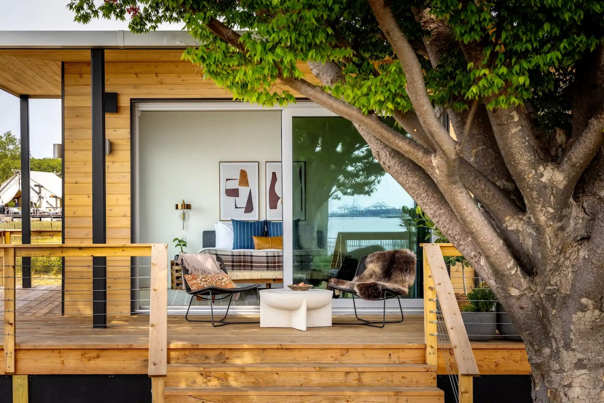 A spacious deck extends from the wooden structure, framed by a large tree. Comfortable seating options, including a stylish lounge chair and a cozy fur throw, complement the outdoor space. Floor-to-ceiling glass doors offer a glimpse into the interior, enhancing the connection with nature.