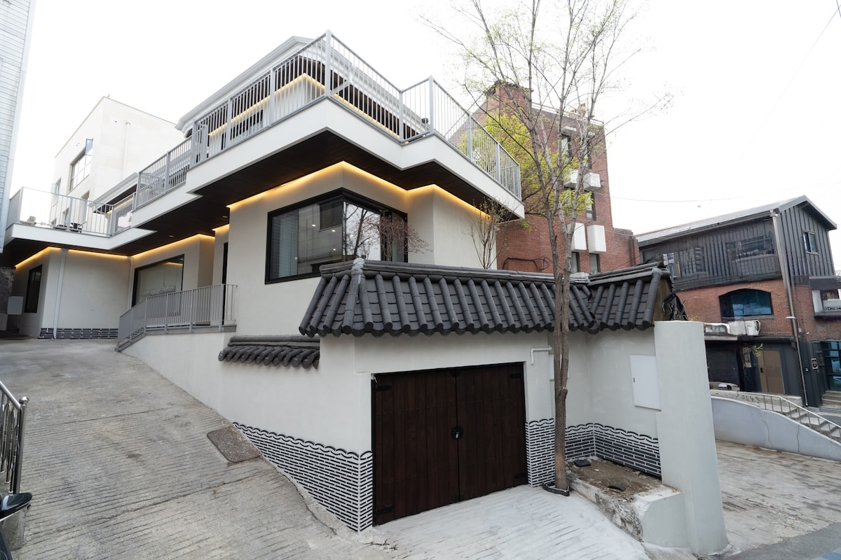 The exterior of a traditional Hanok building features a combination of modern and classic elements. A curved tiled roof contrasts with sleek contemporary design. Soft lighting outlines the balcony and terrace areas, while an upward slope leads to the entrance, framed by a tree on the side.