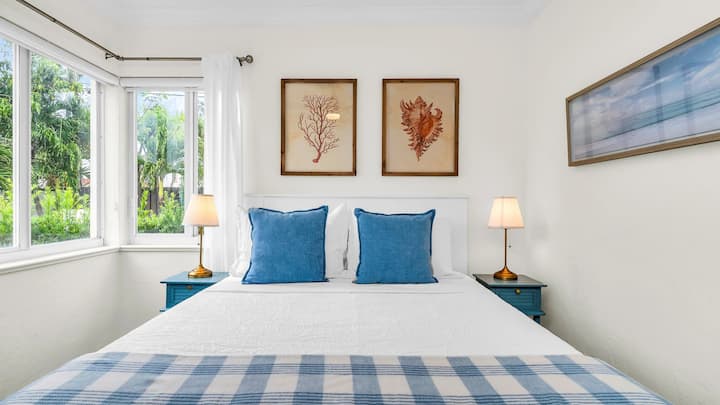 Este sereno tercer dormitorio cuenta con una acogedora cama tamaño queen con sábanas blancas frescas, decoración de inspiración costera y grandes ventanales en las esquinas que llenan el espacio con luz natural y exuberantes vistas al jardín, creando un refugio tranquilo para noches de descanso.