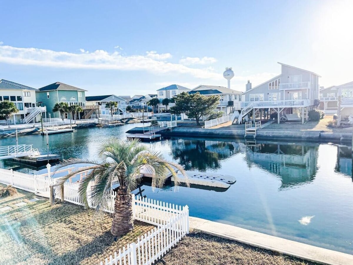 A serene waterfront scene features boats docked along a canal, framed by lush palm trees and a white picket fence. Bright blue skies and reflections on the water create a peaceful atmosphere. Nearby homes are visible, showcasing a mix of architectural styles.